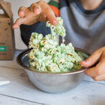 Child's hands mixing green popcorn in a metal bowl on a wooden table.