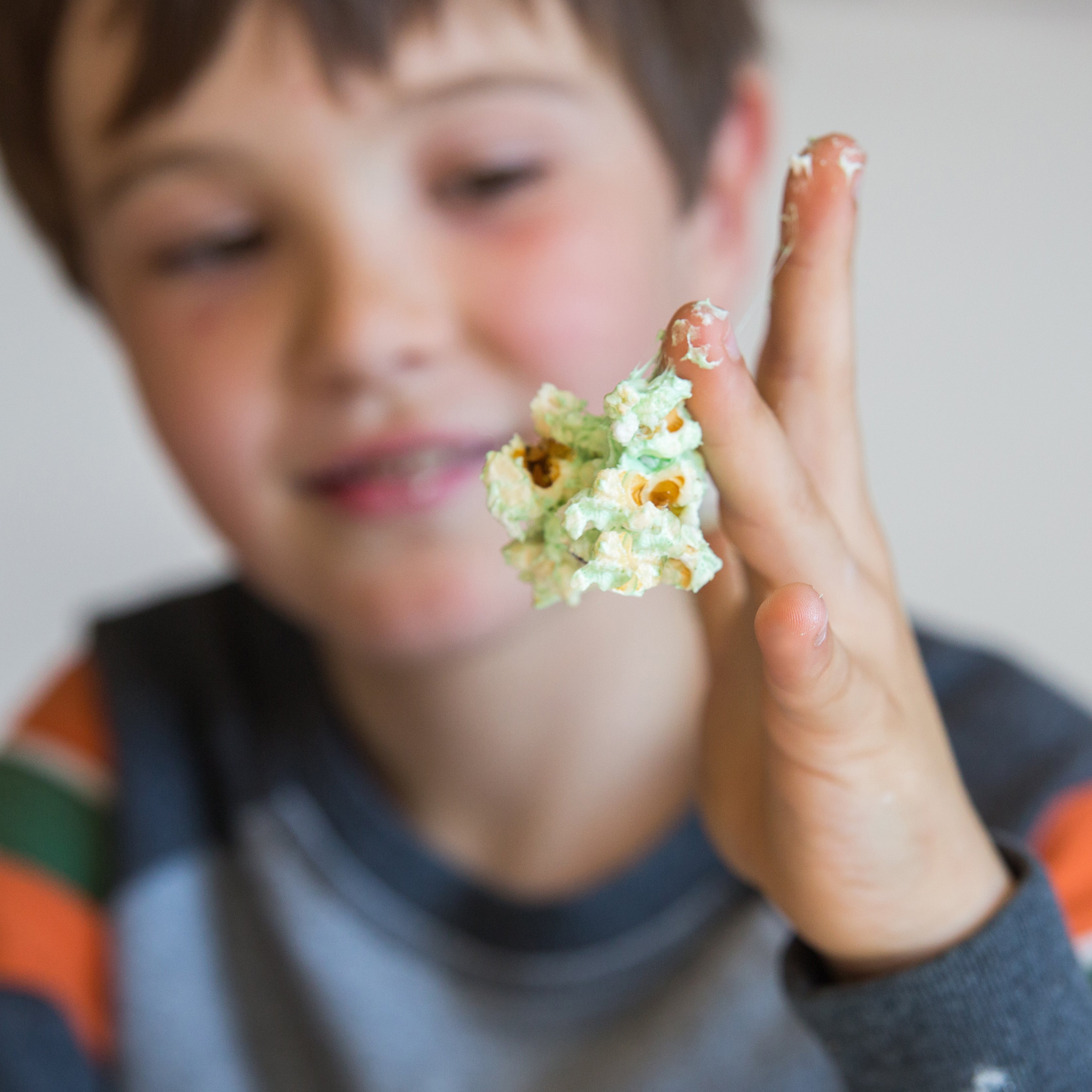 Child holding a piece of popcorn with green coating, smiling, wearing a striped sweater.