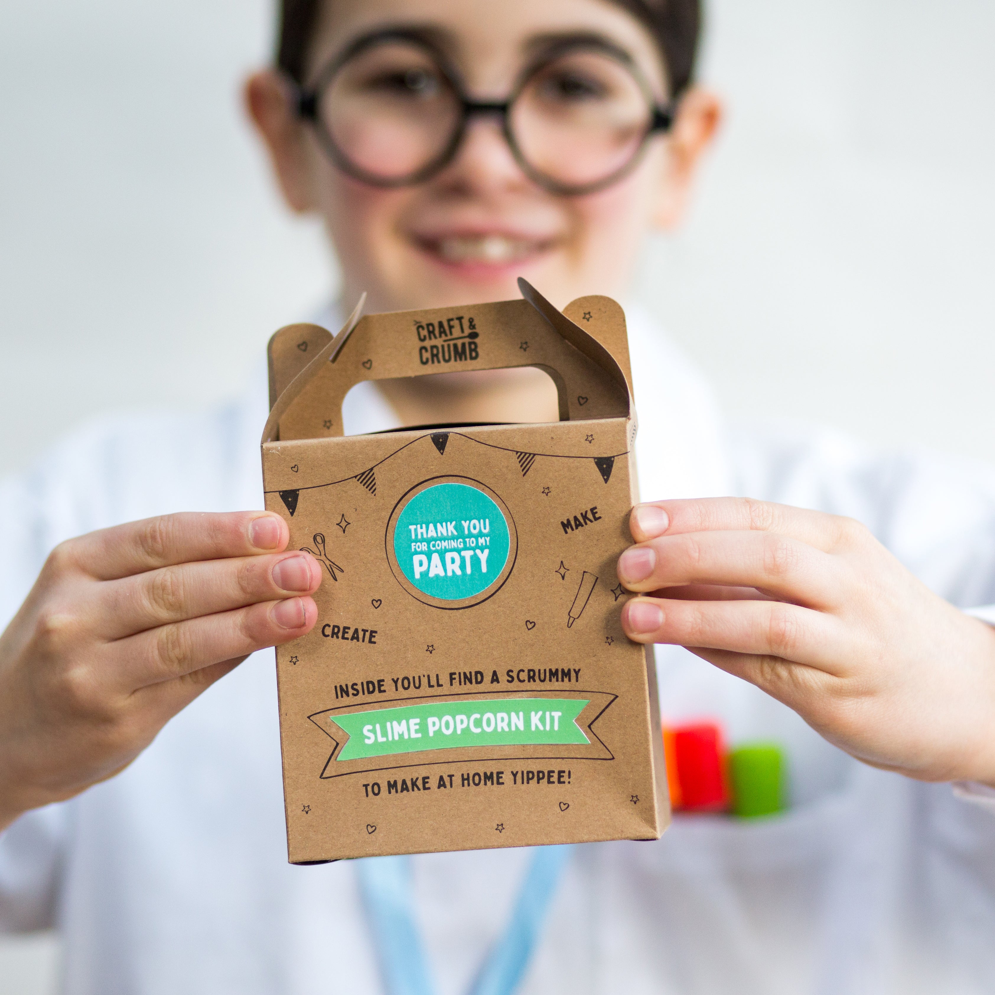 Child holding a "Slime Popcorn Kit" box with a thank you sticker, wearing glasses and a white shirt.